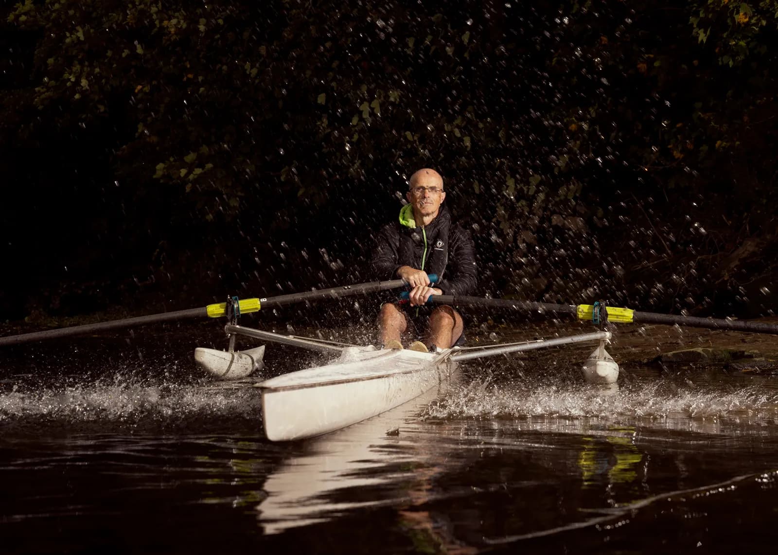 A man in glasses wearing a black jacket with a neon green trim sits in a white rowing scull on dark water, holding an oar, with water splashing around him against a dark background of trees.