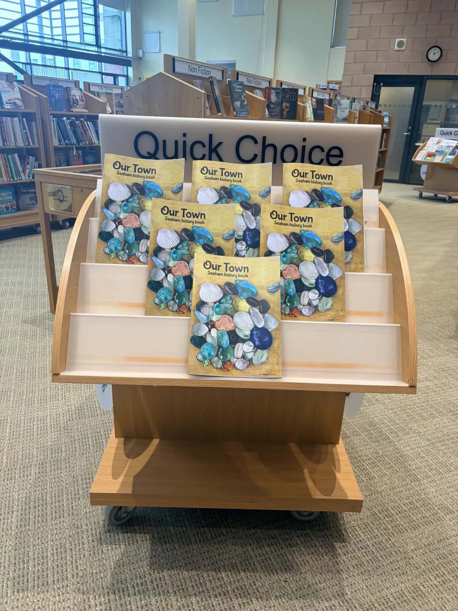 A wooden display stand in a library features several copies of a book titled "Our Town: Seaham history book," with covers depicting colorful pebbles and seashells.