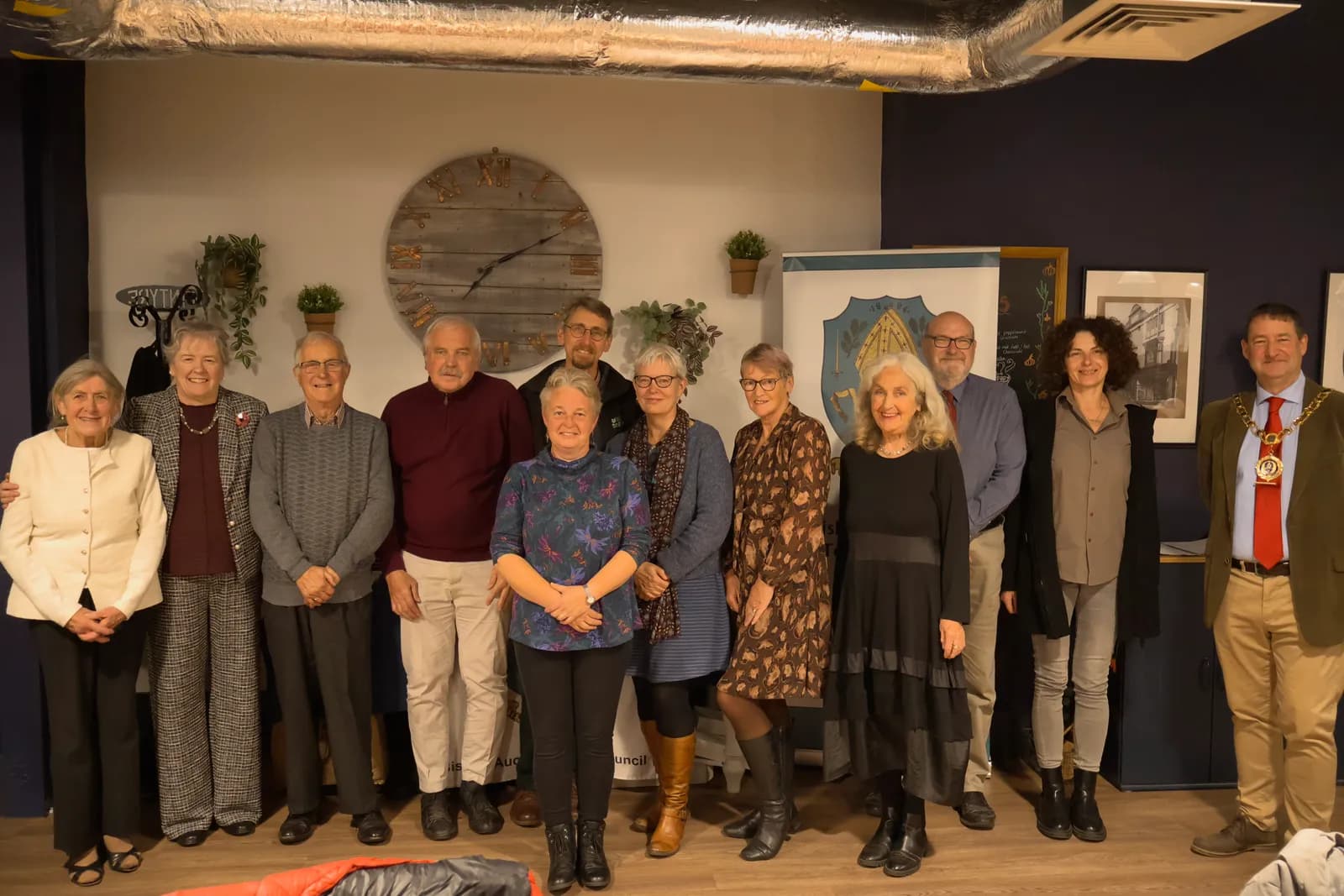A group of thirteen people are posing for a photo in a room with a large wooden clock on the wall.