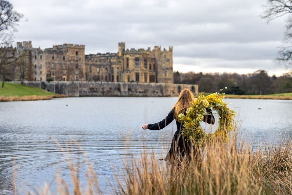 A woman with long red hair, holding a large yellow floral wreath, stands in water with tall grasses in the foreground and a large stone castle across the lake under a cloudy sky.