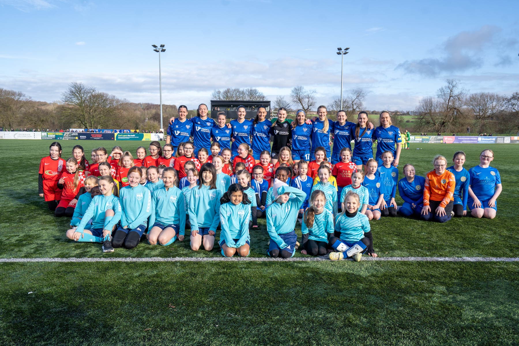 A large group of female athletes of varying ages, including adult women and young girls, pose for a photo on a green sports field under a bright blue sky.