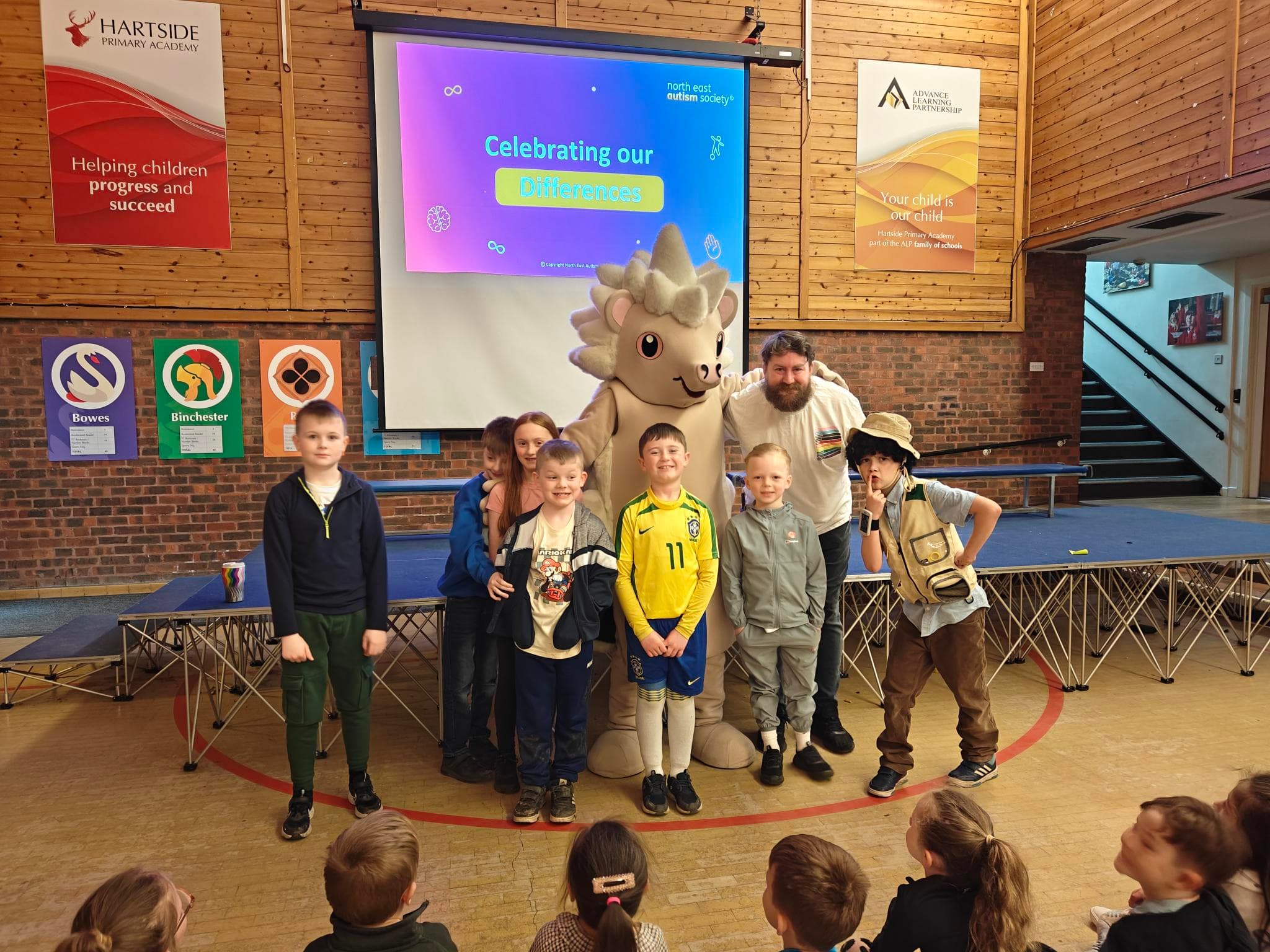 A group of children and adults, including one in a mascot costume, stand on a stage in front of a projection screen that reads "Celebrating our Differences."
