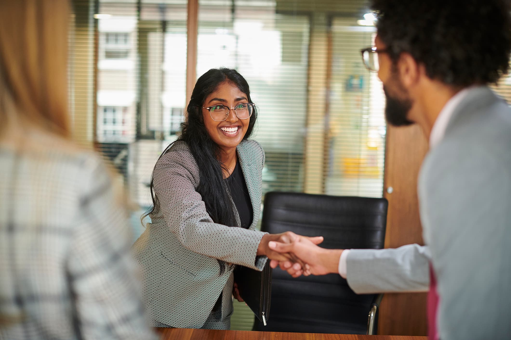 A smiling woman in a gray blazer with long dark hair and glasses shakes hands with a man whose back is partially visible, across a wooden table in an office setting.