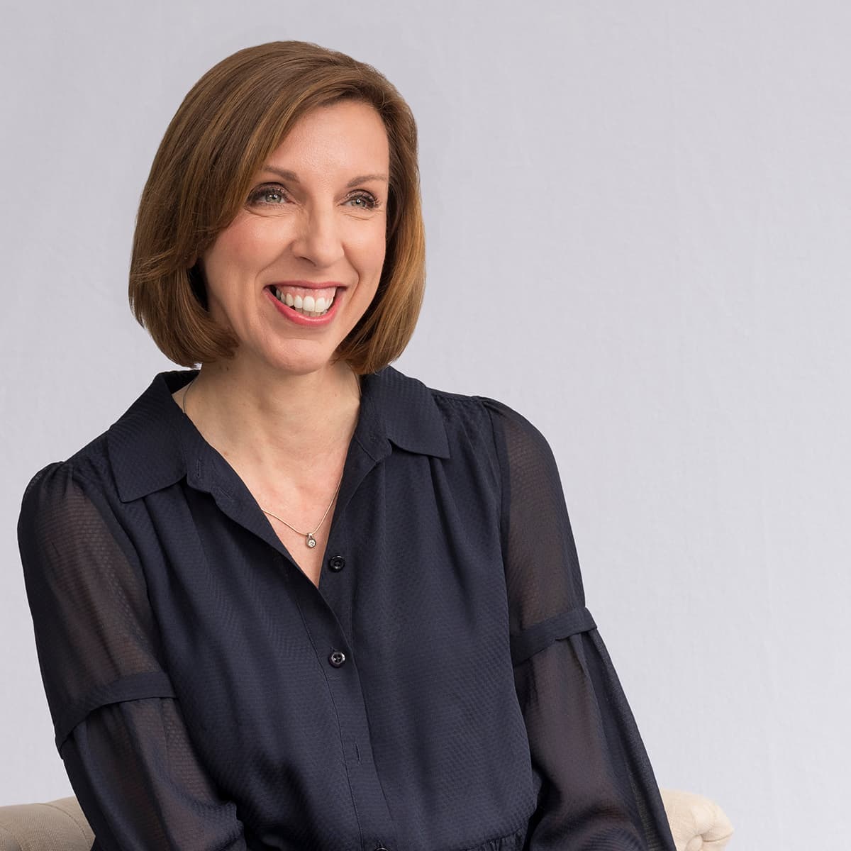 A smiling woman with short brown hair is wearing a dark blue blouse and a delicate necklace with a small pendant, looking to the right of the frame.