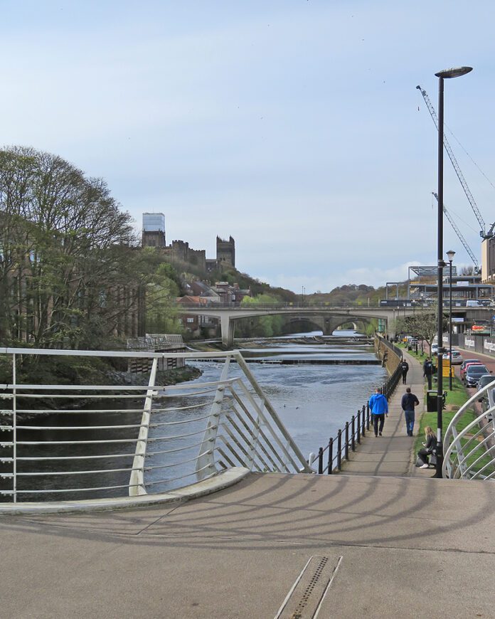 View of Framwellgate Waterside in Durham, looking south from the footbridge towards Milburngate and Framwellgate bridges. Cranes are visible on the right, part of the riverside development, with the central tower of Durham Cathedral in the distance, covered for restoration